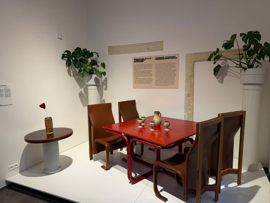 Expozice výstavy HNUSNÝ v Design Museum Prague: červený stůl, hnědé židle, keramika a bílé stojany s rostlinami.